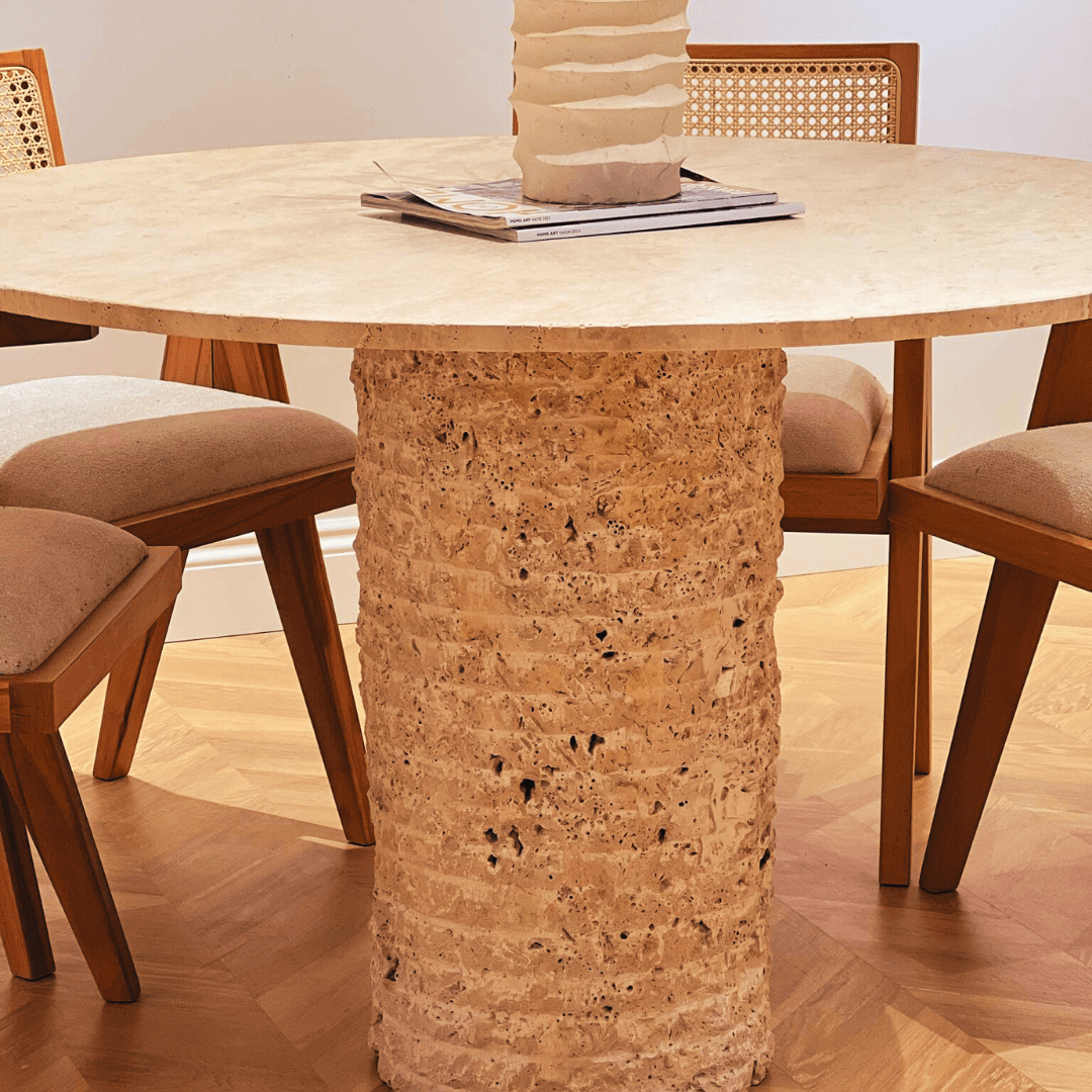The Rustic Round Travertine Dining Table by Travertable, featuring a textured cylindrical base, graces a wooden-floored room. Two chairs with cane backs and beige cushions are partially visible. A white sculptural vase and a magazine add charm atop the table.