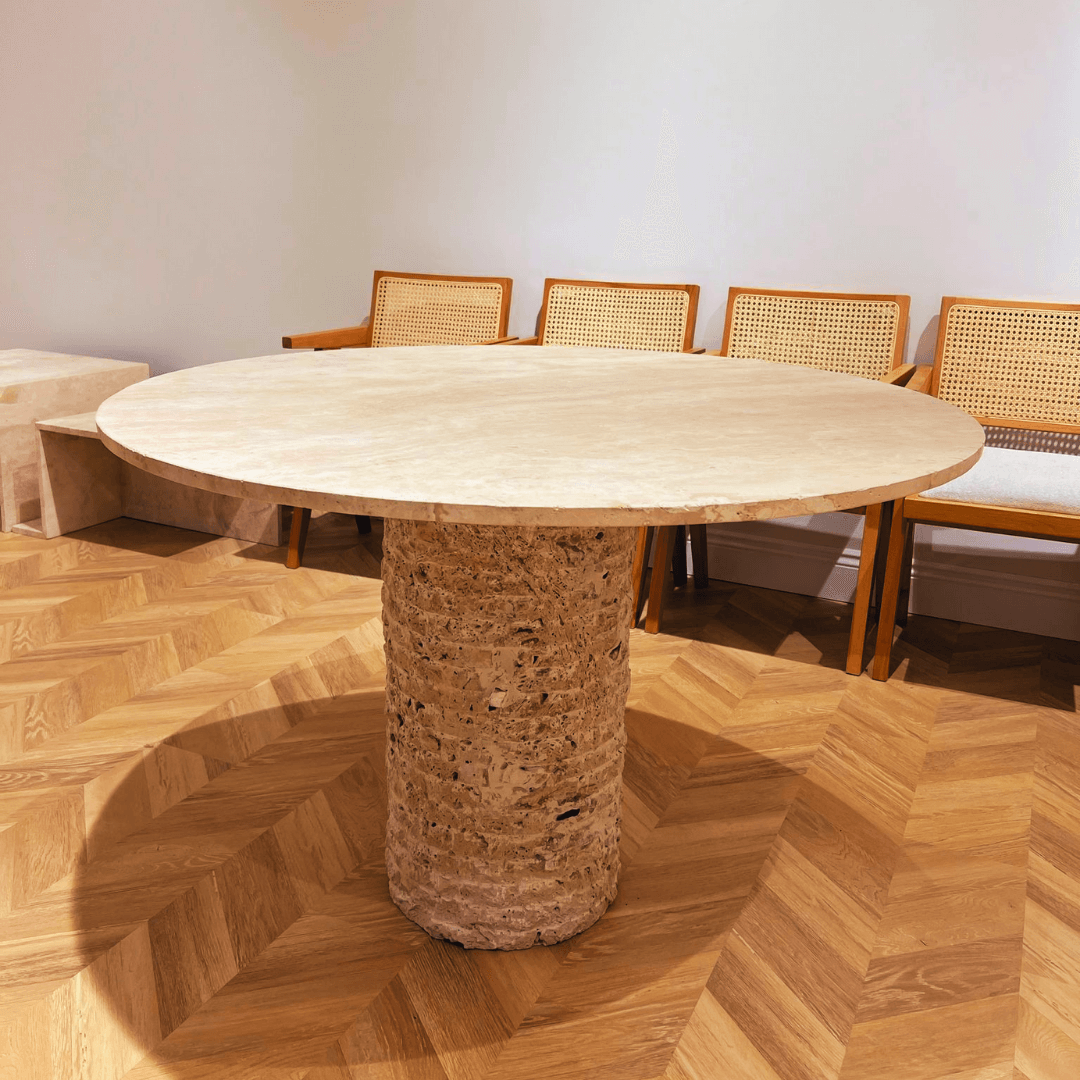 The Travertable Rustic Round Travertine Dining Table, featuring a textured cylindrical base, is placed on a wood-patterned floor. Wooden chairs with woven backrests line the cream-colored wall in the background.