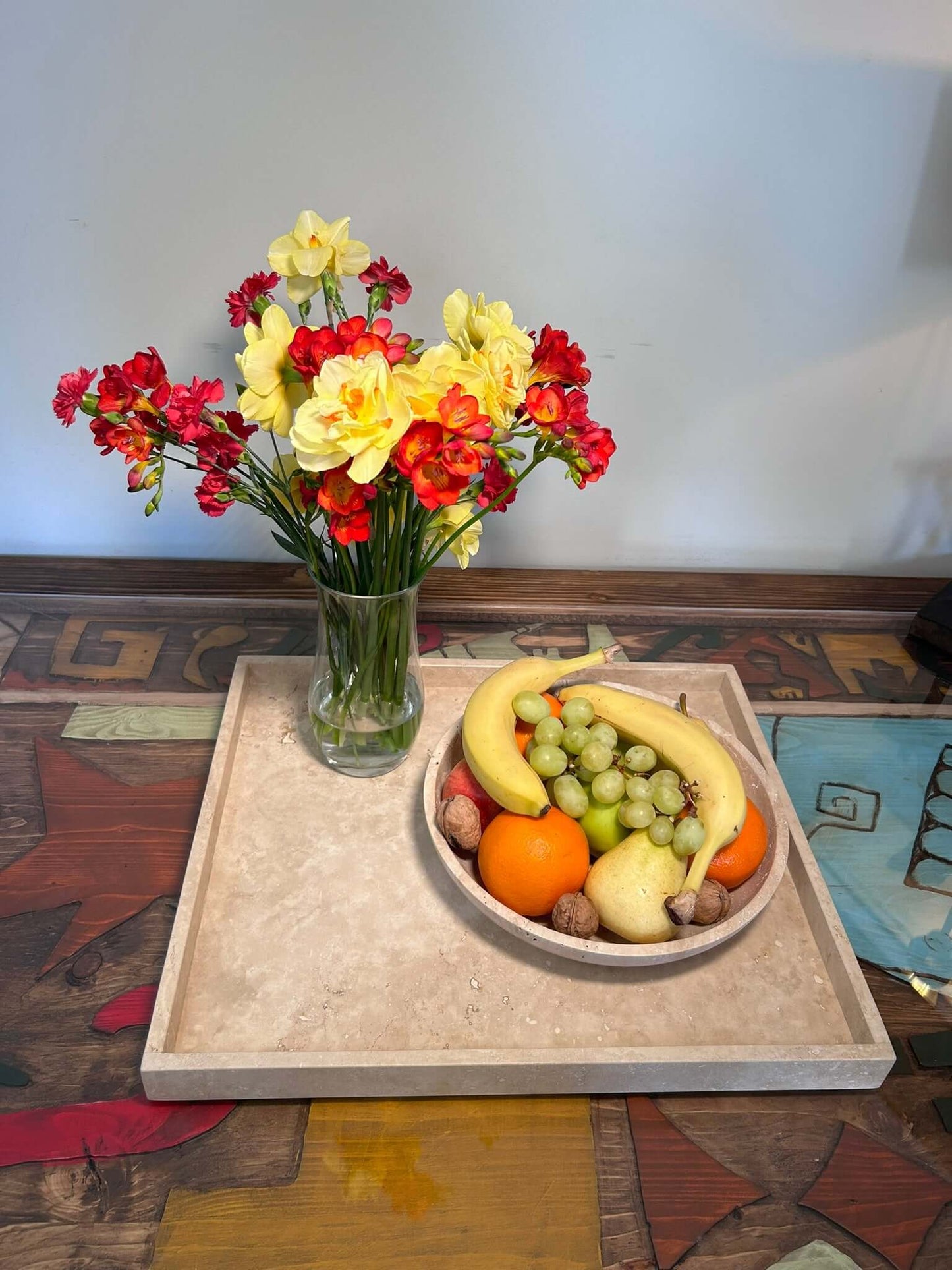 A vase with red and yellow flowers complements a Timeless Travertine Artisanal Bowl by Travertable filled with bananas, green grapes, oranges, pears, and walnuts on a tray. This ensemble sits on a colorful wooden surface that highlights natural artistry in every detail.