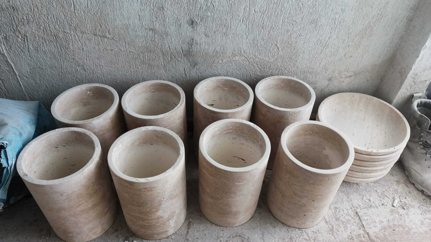 Two rows of cylindrical Golden Sunrise Travertine Vases from Travertable are arranged on a concrete floor near a wall. Nearby, several round stone lids or trays are stacked horizontally, with a blue bag partially visible on the left.