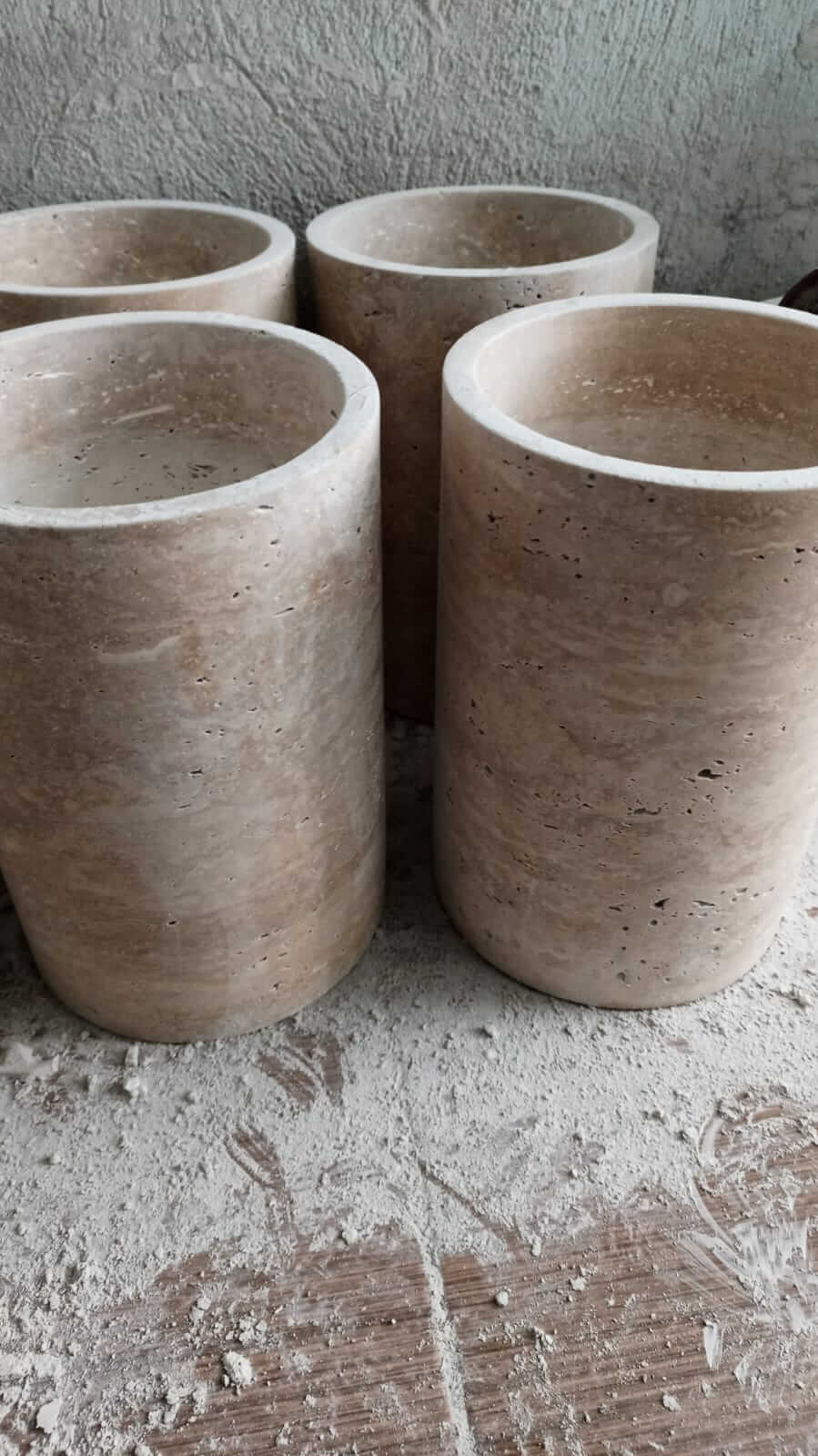 Four Golden Sunrise Travertine Vases by Travertable, featuring a smooth, light brown surface and subtle texture, stand on a dusty surface with a gray, uneven background.