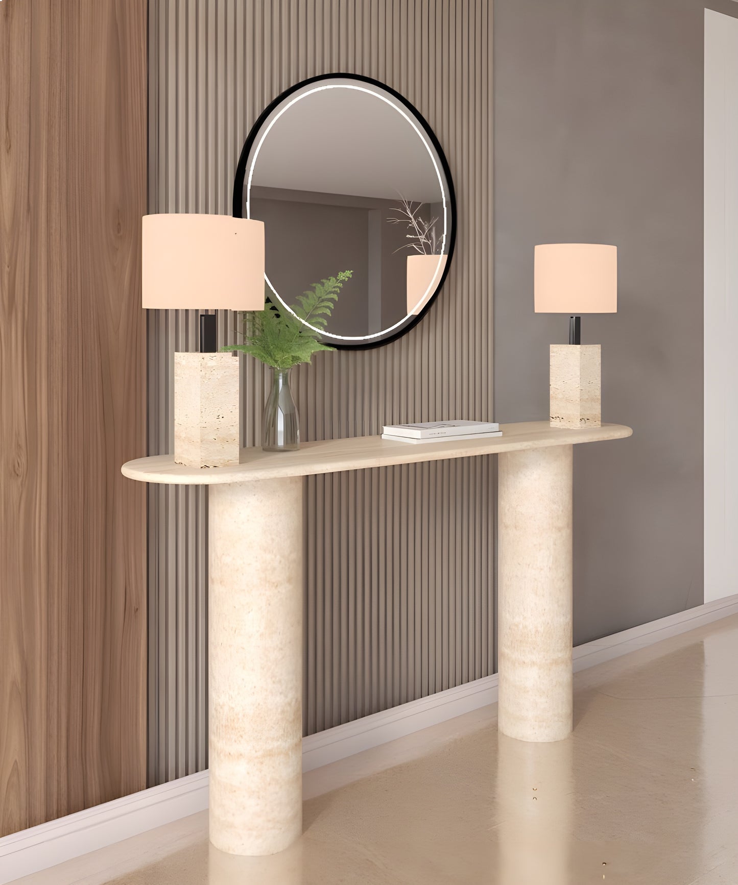 The TRAVERTABLE Ovale Travertine Console Table, with a pure travertine stone base and smooth cylinder design, holds two matching lamps and a glass vase of green foliage. Above, a round black-framed mirror reflects part of the room.