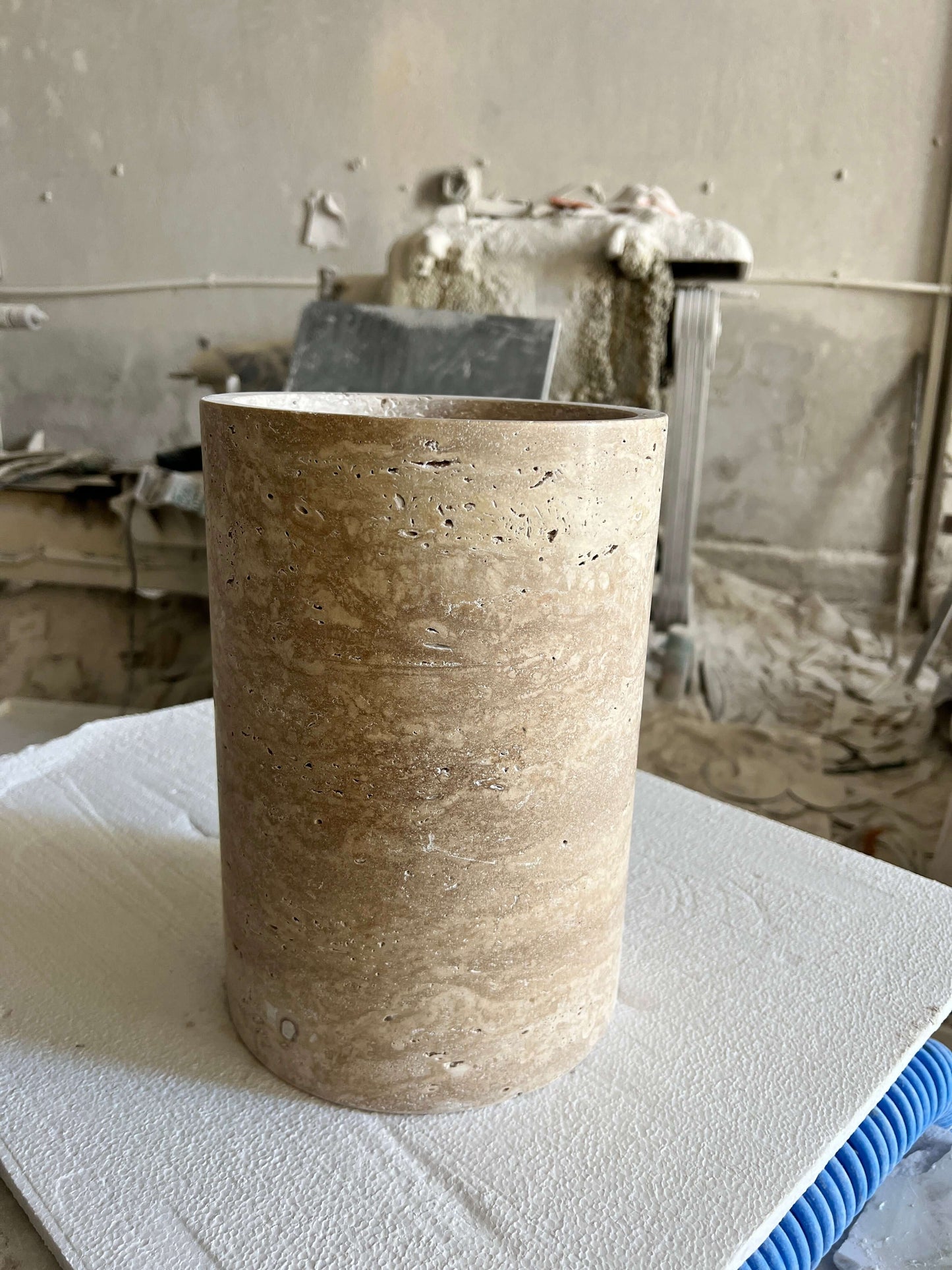 The Golden Sunrise Travertine Vase by Travertable stands on a padded surface in a workshop filled with tools and materials. The walls are unfinished with visible plaster and patches, while dust and debris are scattered around.