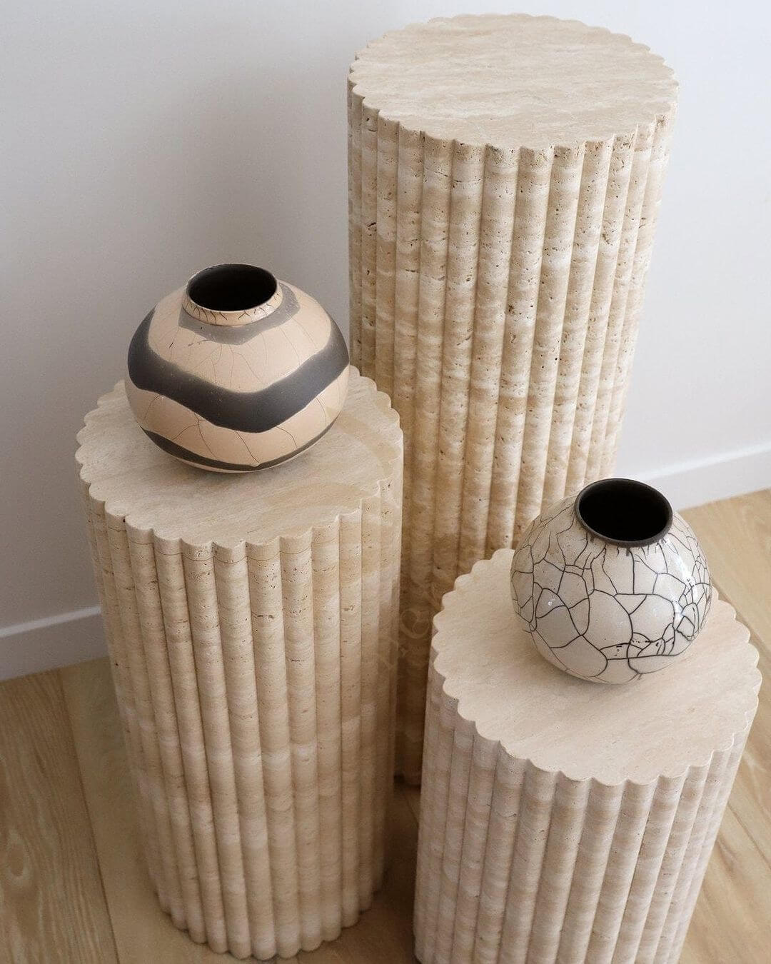 The Travertable Travertine Elegance Pillar Trio, a sculptural nested set with fluted textures and varying heights, displays two small round vases on a wooden floor—one patterned in beige and gray and the other featuring a cracked beige design.