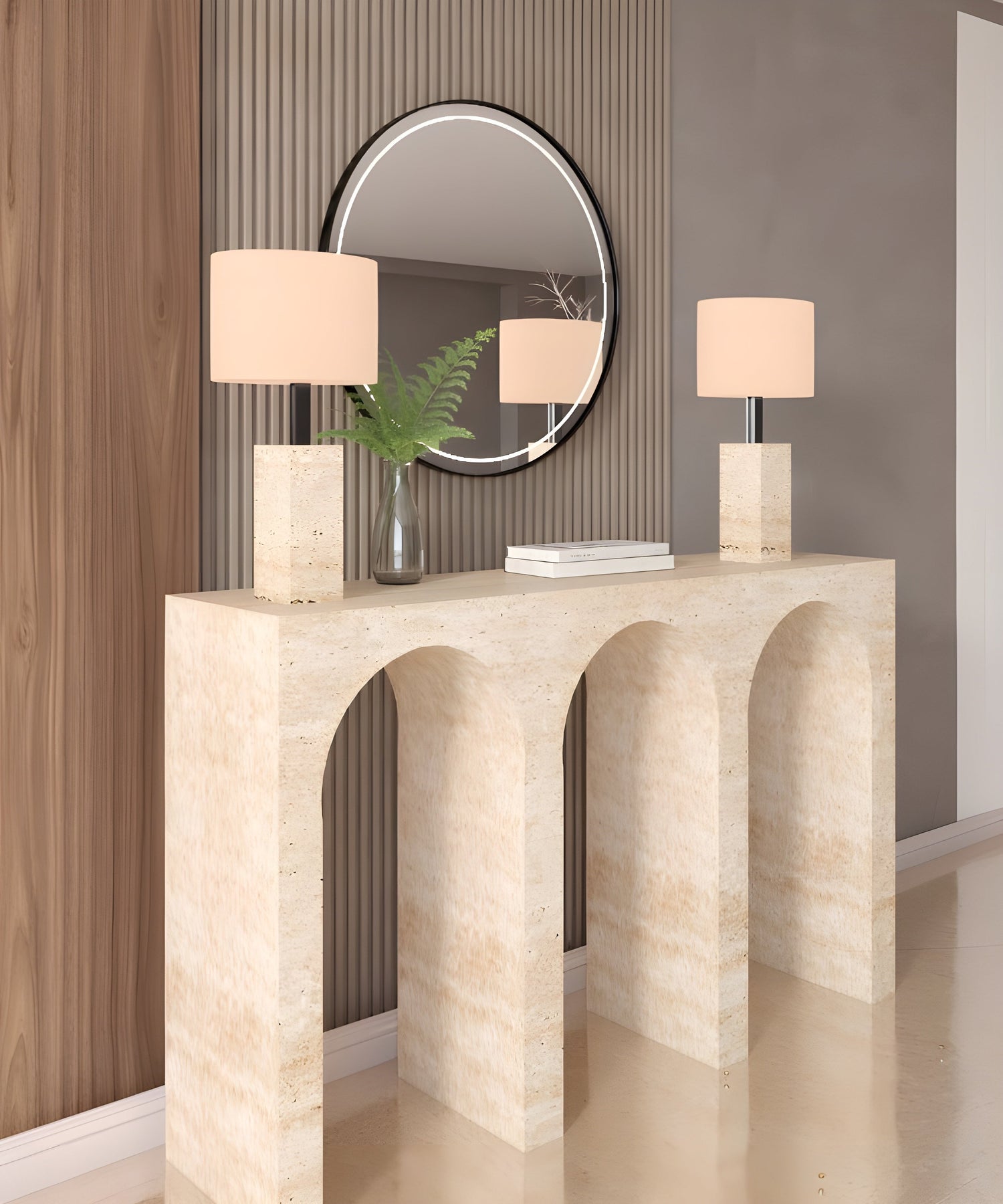 A modern entryway with a beige stone console table featuring three arches, two matching lamps with beige shades, a glass vase with green fern leaves, books, and a round mirror on the wall.