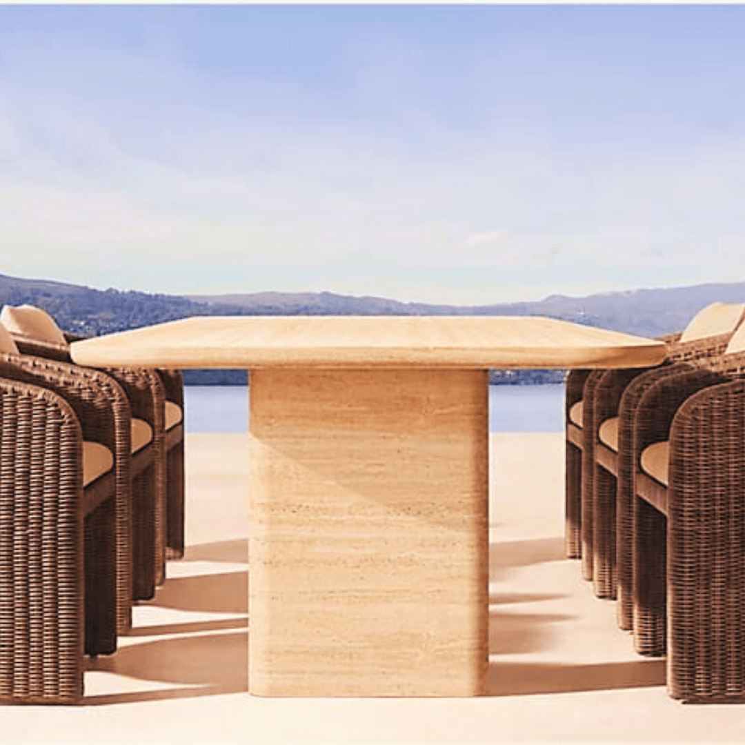 Wicker chairs encircle the Travertable Majestic Travertine Dining Table, featuring customizable tabletop thickness, on a sunlit patio. A clear blue sky and distant mountains enhance the modern design setting.
