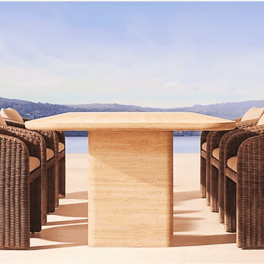 Wicker chairs encircle the Travertable Majestic Travertine Dining Table, featuring customizable tabletop thickness, on a sunlit patio. A clear blue sky and distant mountains enhance the modern design setting.
