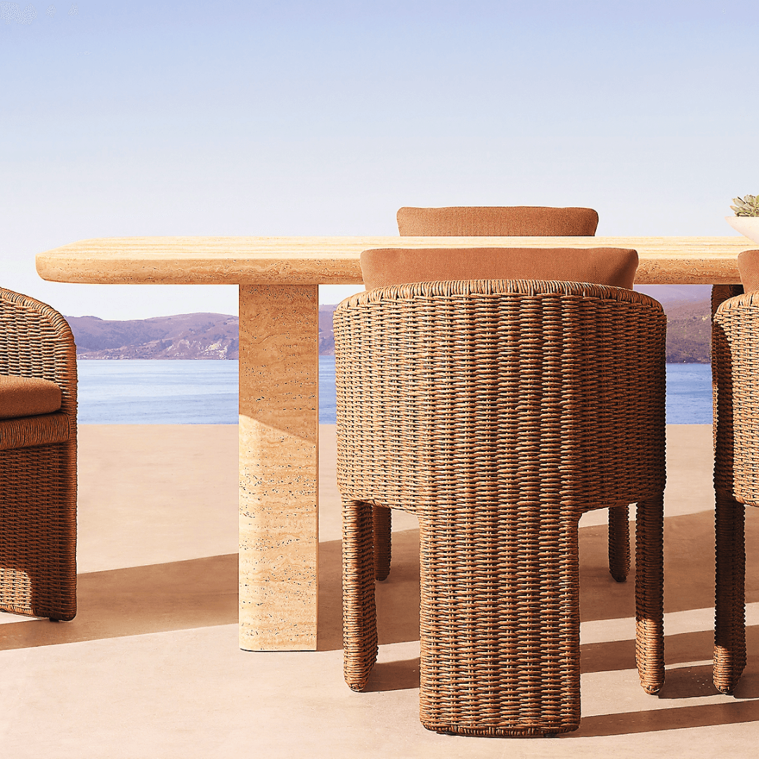 Brown-cushioned wicker chairs encircle The Majestic Travertine Dining Table by Travertable, providing a modern aesthetic. With customizable tabletop thickness, this patio setup overlooks a clear blue lake and distant hills under a perfect sky.