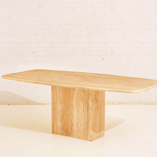 A minimalist rectangular wooden table with a slab top and block base stands against a light brick wall and floor, reminiscent of Travertables Serenity Travertine Dining Table. Its elegant form mirrors the premium travertines beauty, casting a faint shadow to the left.