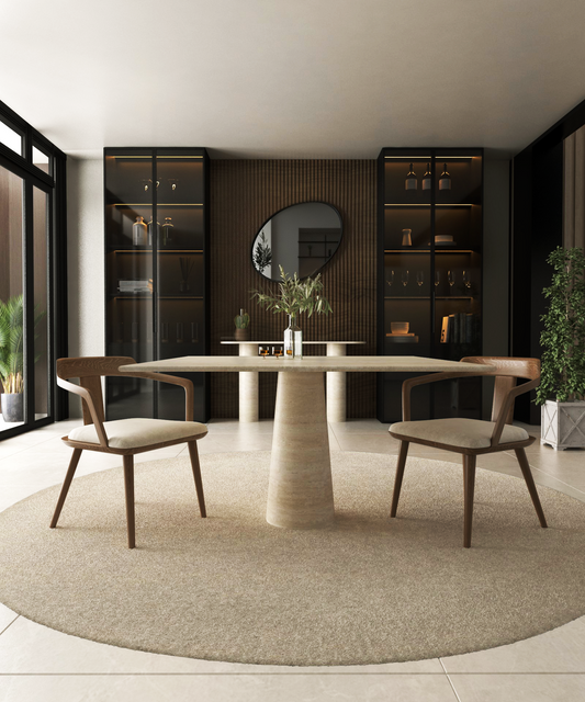 A modern dining room featuring the TRAVERTABLE Square Travertine Dining Table with Conical Pedestal Base, two wooden chairs, a circular rug, built-in shelves with decor, a round mirror on a wood-paneled wall, large windows, and green plants.