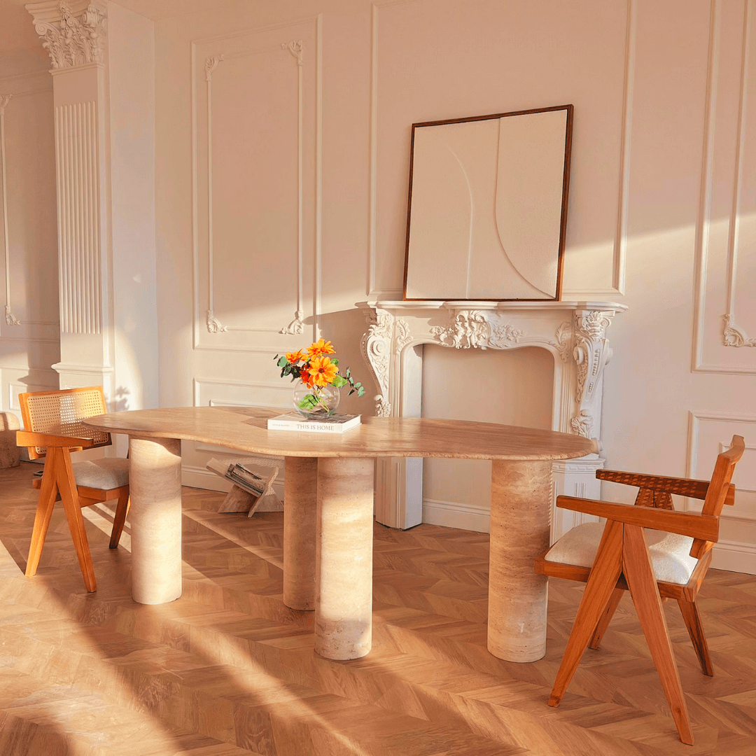 A sunlit dining room exudes natural elegance with Travertable’s Cascade Travertine Dining and Meeting Table and two wooden chairs, centered by a bouquet of flowers. Elegant white molding, a decorative mantle, simple framed artwork, and warm light enhance its cozy ambiance.