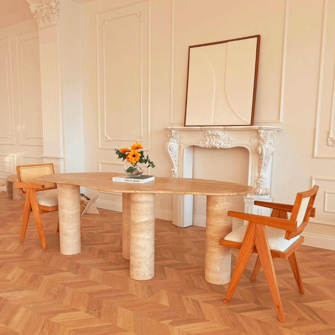 A chic interior with the Travertable Cascade Travertine Table featuring customizable options, paired with two modern wooden chairs with white cushions. A vase of vibrant flowers and a book enhance the natural elegance against elegant white wall moldings.