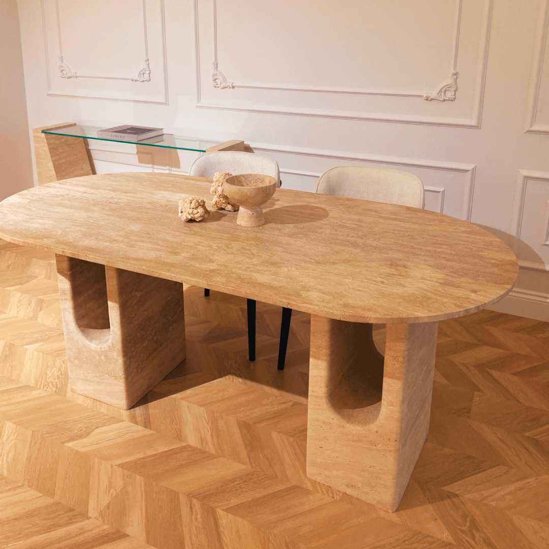 A modern dining room features The Regal Arch Travertine Dining and Meeting Table by Travertable as a stunning centerpiece. This large oval stone table with thick legs is paired with a matching stone bowl, two upholstered chairs, and accented by herringbone wood flooring and a decorative white wall.