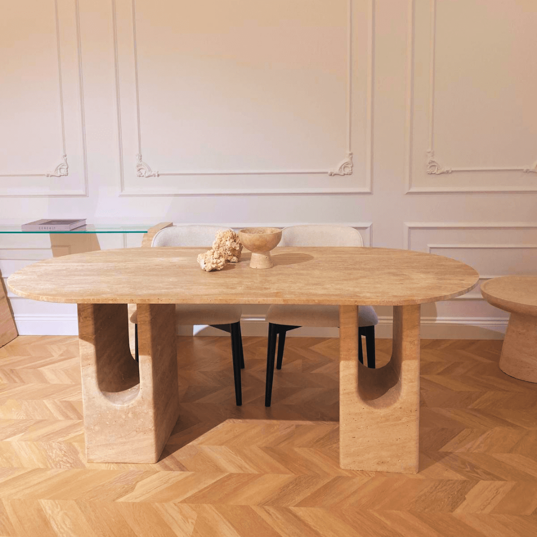 A modern dining room centers on Travertables The Regal Arch Travertine Dining and Meeting Table with distinct curved supports, adorned with a decorative bowl and dried flowers. Its complemented by two black chairs, wooden herringbone flooring, and pristine white walls.