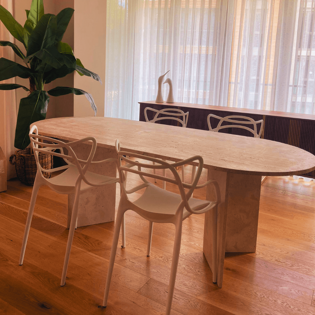 A modern dining room showcases The V-Leg Travertine Dining Table by Travertable, featuring an oval travertine top and elegant V-shaped legs. Its paired with intricately designed white chairs on a wooden floor, accented by a large plant in the corner and semi-transparent curtains that bathe the room in natural light.