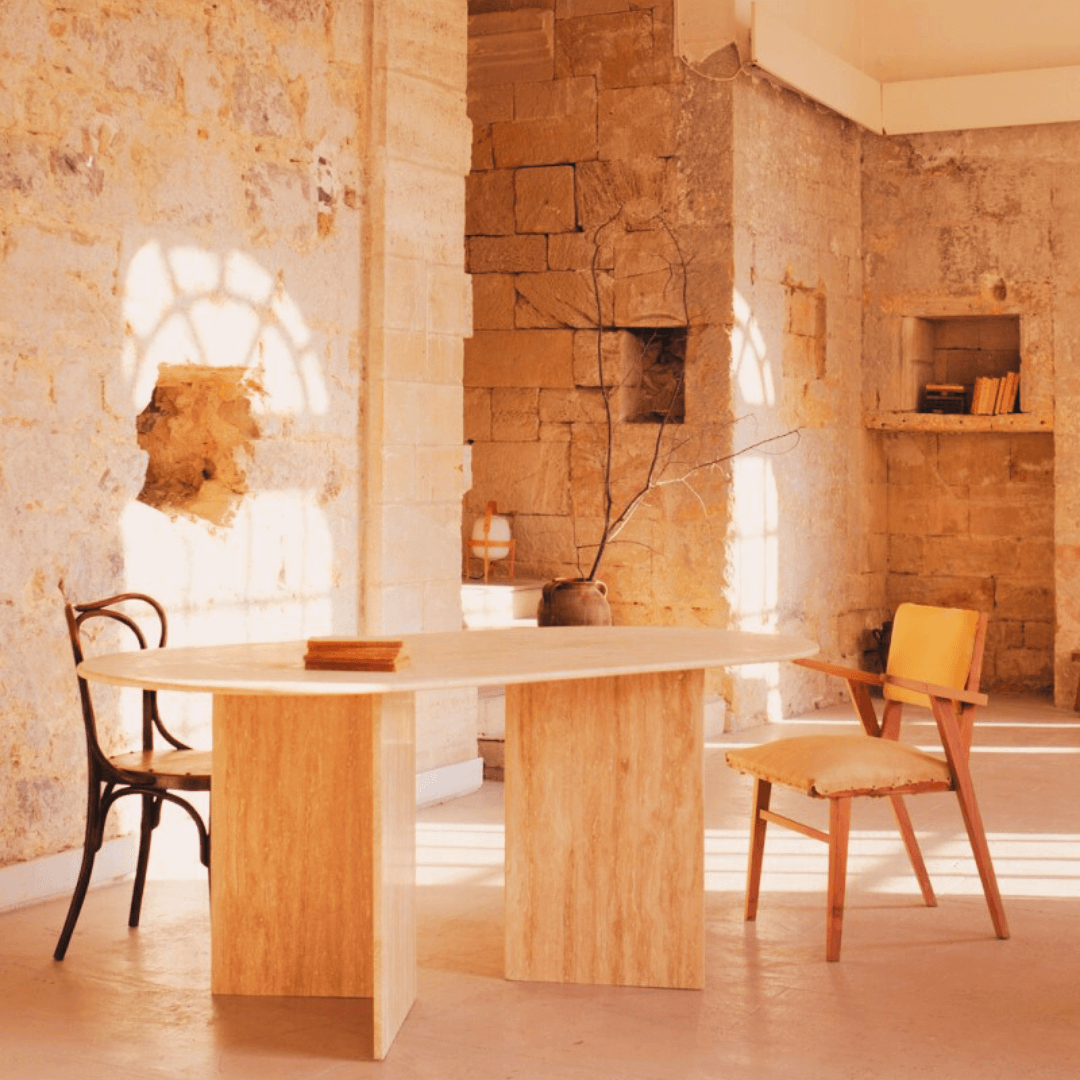 In a rustic room with textured stone walls, Travertables V-Leg Travertine Dining Table stands out, flanked by two chairs—one dark and one light with yellow padding. A vase holding a bare branch sits near an open book on the table, as sunlight filters through the window.