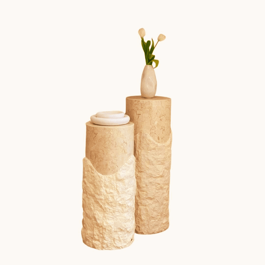 Two Rustic Charm Travertine Pedestals from Travertable, reminiscent of travertine columns with a natural texture, hold a decorative vase of small white flowers. The vases earthy appearance contrasts beautifully with the smooth white blooms against a plain, neutral background.
