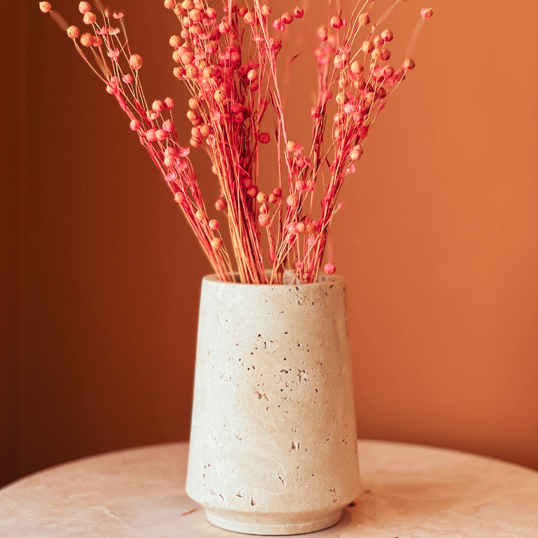 A Classic Beige Travertine Vase by Travertable holds dried orange-red flowers with small round buds on a marble surface. The warm, orange-brown wall enhances the minimalist, cozy atmosphere.