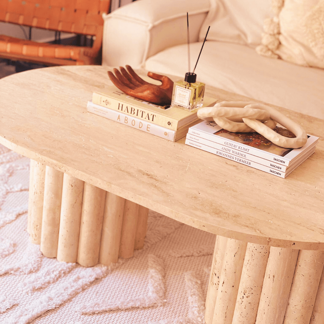The Handcrafted Italian Travertine Coffee Table by Travertable, with its beige oval surface and cylindrical legs, showcases fine craftsmanship. It displays books, a wooden hand sculpture, a reed diffuser, and twisted candles on a textured white rug with a sofa and leather chair nearby.