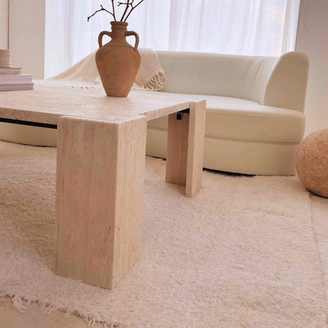 A minimalist living room includes a white curved sofa, a large beige rug, and Travertables Modern Rustic Travertine Coffee Table. A clay vase with branches adorns the table, while sheer curtains filter light to enhance the serene atmosphere.