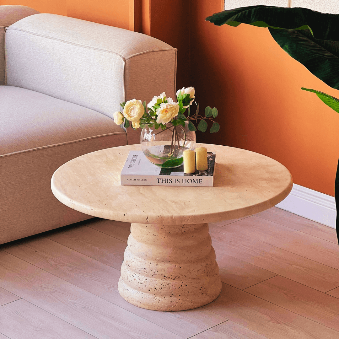 A Designer Travertine Marble Coffee Table by Travertable, in beige with a sculpted base, displays a small vase of yellow roses, the book This is Home, and two yellow candles. It sits on light wood flooring beside a cream sofa against an orange wall, with a large plant adding natural beauty to the scene.