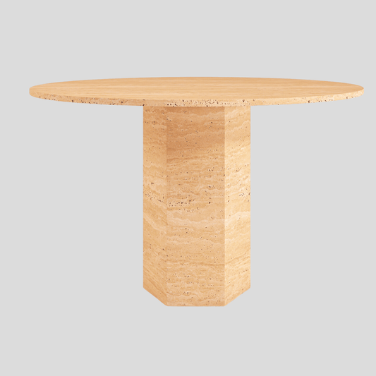 The Travertable Hexagon Round Travertine Dining Table features a round beige marble top on a solid hexagonal travertine base, set against a plain gray backdrop.