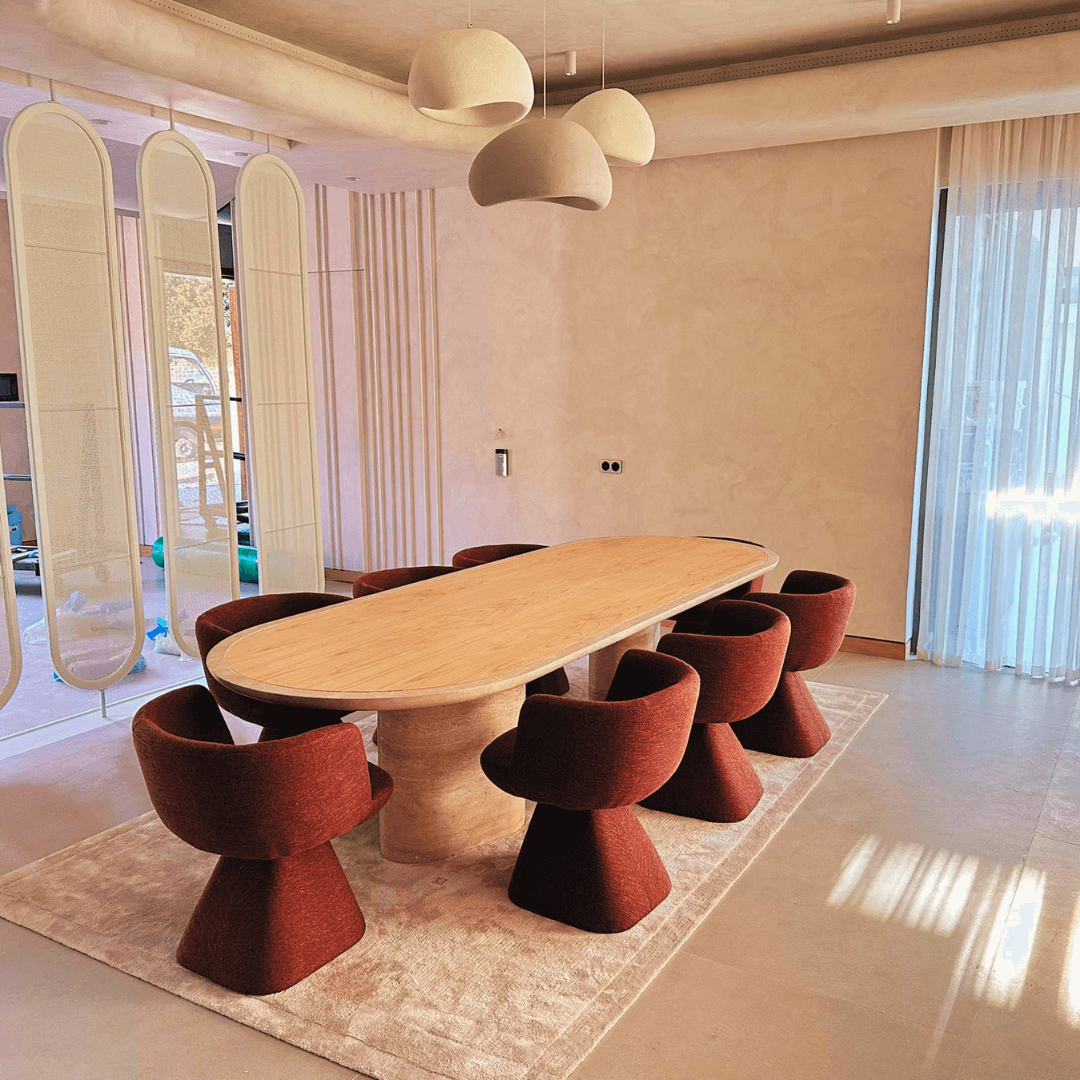 A modern dining room showcases The Harmony Travertine and Walnut Dining Table by Travertable, surrounded by six red chairs on a light rug. Three artistic pendant lights illuminate the area, set against soft beige walls with sheer curtains and three tall mirrors adorning one wall.