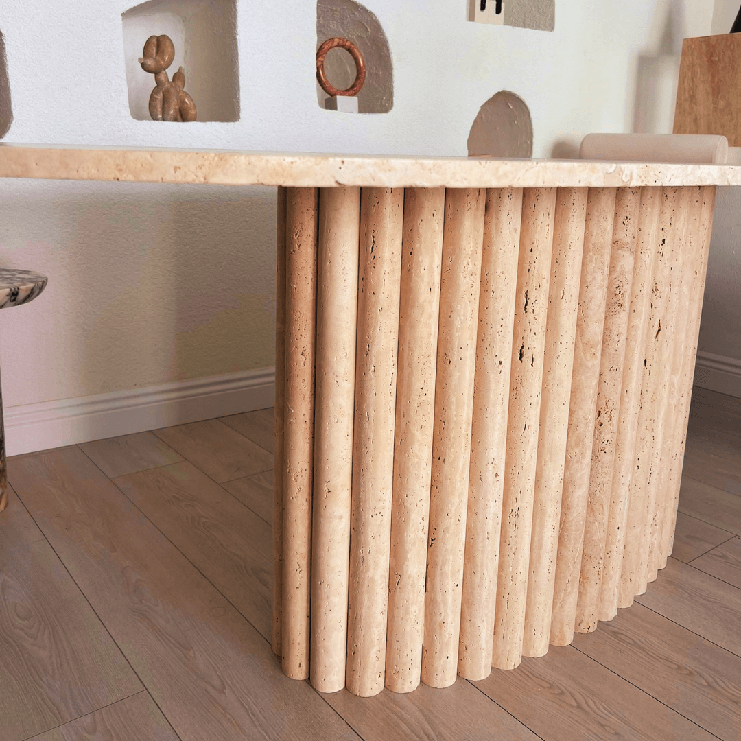 The Elysian Travertine Feast Table by Travertable features a beige travertine top with a ribbed pedestal base, elegantly positioned on a light wooden floor. The backdrop includes a white wall with recessed shelves holding decorative items.