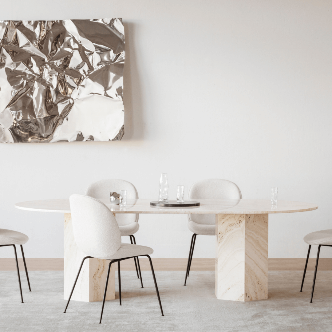 A modern dining room showcases The Hexaform Travertine Dining Table by Travertable, with a round top and hexagonal legs, accompanied by five white chairs. A metallic abstract artwork decorates the wall, clear glass vases rest on the customizable surface, and a light grey rug covers the floor.