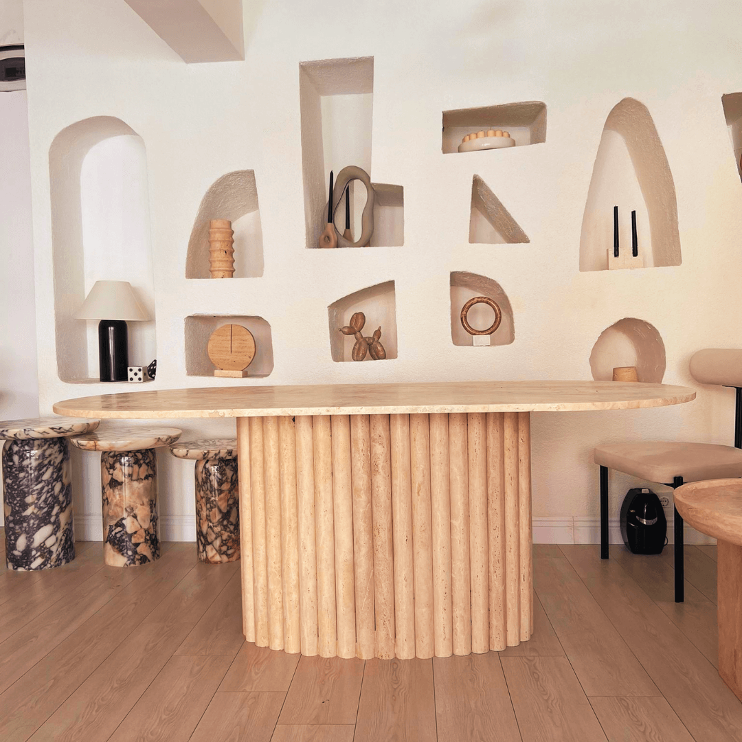 A modern interior showcases the Elysian Travertine Feast Table by Travertable, featuring a round wooden top and ribbed pedestal. Decorative items fill shelves in the white textured wall. Marbled stools and a beige bench provide seating, completing the minimalist and artistic room with subtle customization options.