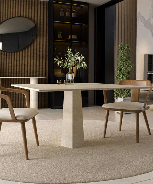 A modern dining area showcases the TRAVERTABLE Square Travertine Dining Table with Rockcone Center Base, two cushioned wooden chairs, a greenery-filled vase, and a round beige rug. A dark shelf, round mirror, and potted plant accent the background.