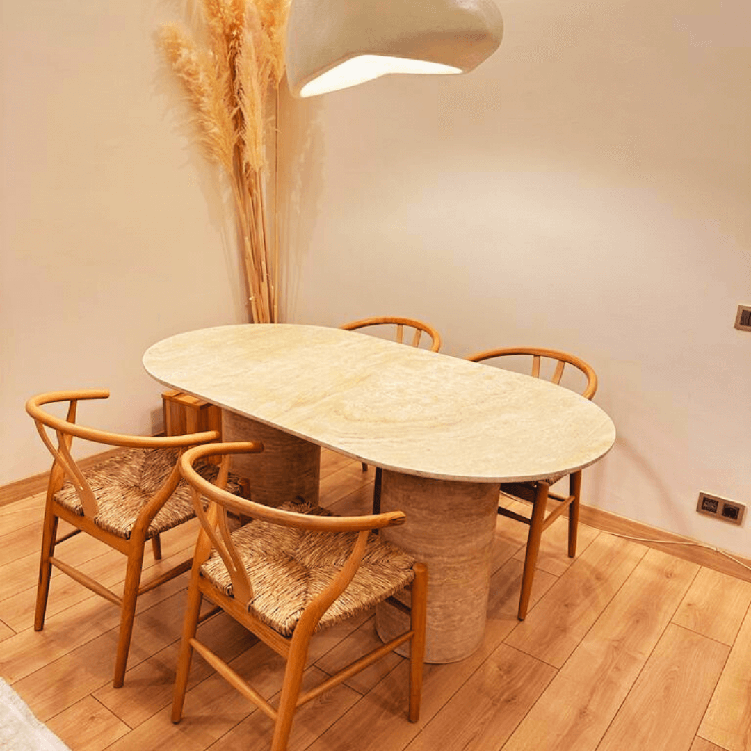 A contemporary dining room highlights The Monarch Travertine Dining Table by Travertable, paired with four wooden chairs with woven seats on a light wood floor. A large hanging lamp is above the table, and tall dried plants adorn a corner by the wall.