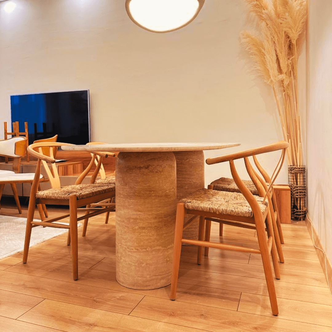A contemporary dining room features The Monarch Travertine Dining Table by Travertable with four woven-seat chairs on a light wood floor. A flat-screen TV is wall-mounted, and a tall vase of pampas grass in the corner enhances the natural stone charm.