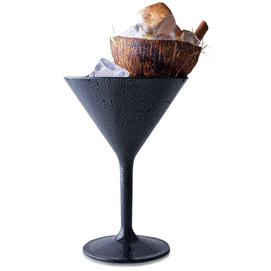 A Mamaco Home black Large Martini Glass, made from shatterproof polycarbonate, holds ice cubes topped with a coconut shell filled with more ice and crowned by a cinnamon stick, while glistening with condensation.