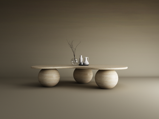 The TRAVERTABLE Wavy Travertine Coffee Table features an irregular oval top and three stone ball bases. Styled with a glass vase, branch, and ceramic pitchers on a tray, it sits against a seamless gradient of neutral beige tones.