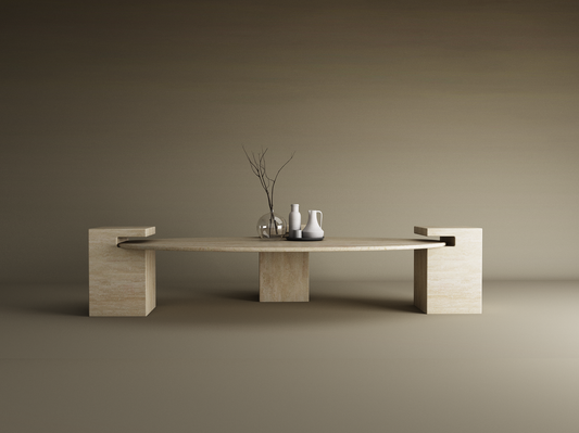 The TRAVERTABLE Ellipse Travertine Coffee Table with Square Base in Timeless Travertine features a stone top, three rectangular legs, and two side cut-outs. Styled with a glass vase of branches, white pitcher, and dark cup on a plain beige background.