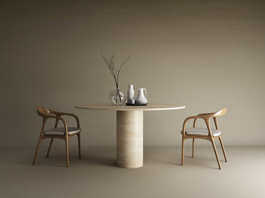 The TRAVERTABLE Oval Travertine Dining Table with a cylindrical center base is styled with minimalist vases and twigs. Two wooden chairs with curved backs and white cushions flank the pure travertine stone table in this serene dining area.