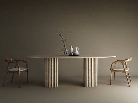 The Ellips Travertine Dining Table by TRAVERTABLE, featuring a fluted solid stone base, is styled with minimalist ceramics and a branch in a glass vase. Two wooden chairs with white cushions face the table against a neutral beige backdrop.