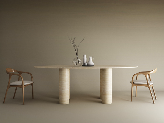 A minimalist dining setup features TRAVERTABLEs Ellips Travertine Dining Table with Cylindrical Base, two wooden chairs with white cushions, and ceramic vases with bare branches, all set against a neutral beige background.