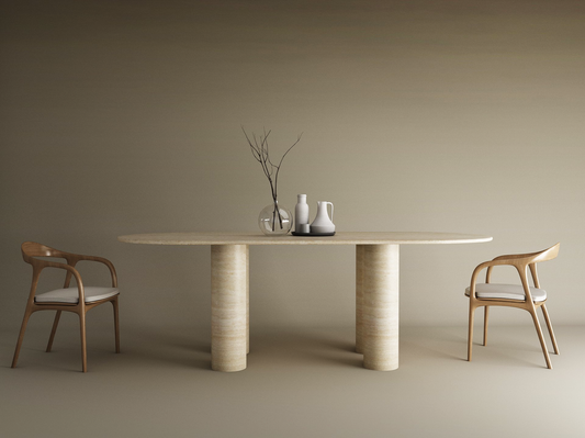 A minimalist dining area features the TRAVERTABLE Oval Travertine Dining Table with Cylindrical Base, two wooden chairs with white cushions, and a glass vase centerpiece with branches and ceramic jars set against a neutral backdrop.