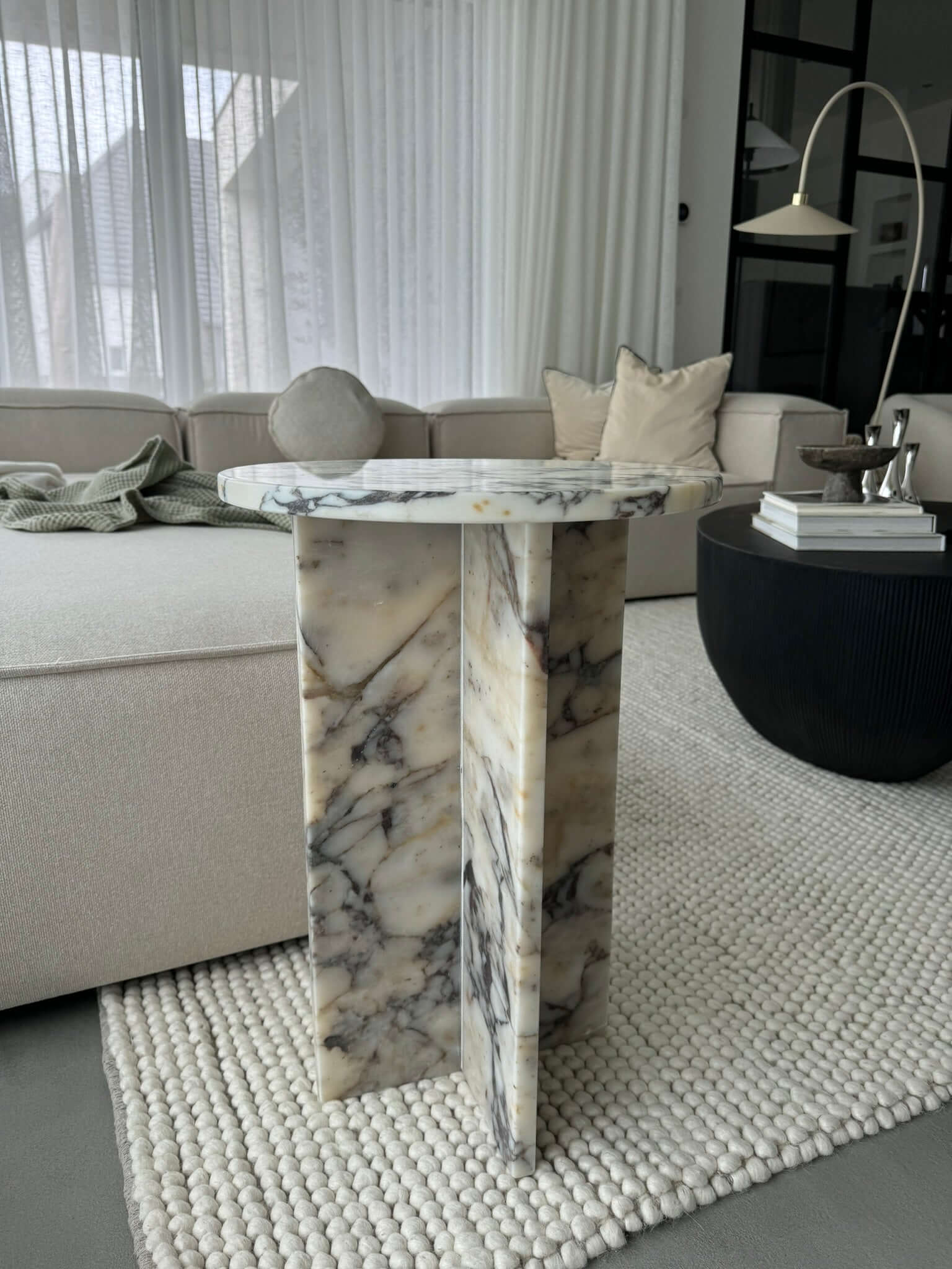 A modern living room displays a stunning Travertable Ela Prestige Calacatta Viola Marble coffee table with a round top and geometric base. A beige sofa with pillows and a knitted blanket complements the white rug, while a black coffee table holds books and decor.