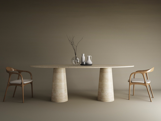 The TRAVERTABLE Ellips Travertine Dining Table with two conical stone bases stands amid wooden chairs with white cushions. Minimalist vases, bowls, and a glass branch vase sit atop the natural tabletop; the background is neutral.