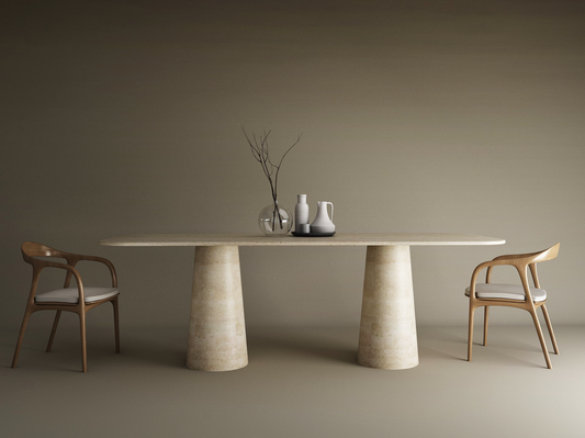 A minimalist dining setup features the TRAVERTABLE Rectangular Travertine Dining Table with a conical pedestal organic stone base, paired with two wooden chairs with white cushions. Simple decor—a vase with branches and two pitchers—accents a plain taupe backdrop.