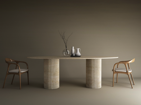 A minimalist dining setup with the TRAVERTABLE Ellips Travertine Dining Table featuring a vertical groove organic stone base, two wooden chairs with white cushions, and vases with branches as a centerpiece on a neutral-toned background.