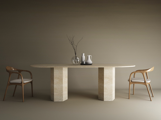 A minimal dining setup features the TRAVERTABLE Oval Travertine Dining Table with a hexagonal stone base, two wooden chairs with white cushions, three ceramic vessels, and a glass vase with branches, all set against a plain brown background.