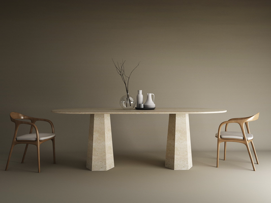 A minimalist dining setup features the TRAVERTABLE Oval Travertine Dining Table with Tapered Round Base, two wooden chairs with white cushions, a vase with dried branches, and two pitchers, all set against a plain beige background.