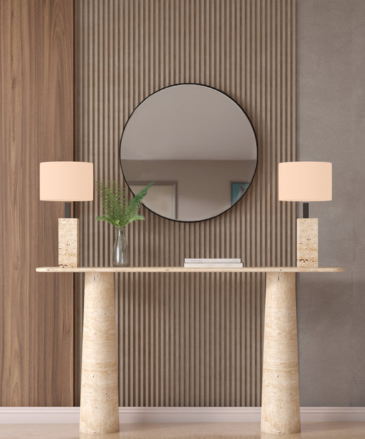 A modern entryway features a round mirror on a ribbed wood wall, the TRAVERTABLE Ovale Travertine Console Table with conical pedestal base, two beige rectangular-shade lamps, and a glass vase filled with green fern leaves.