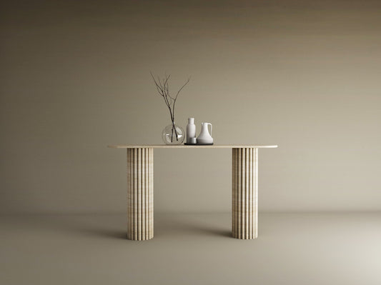 A TRAVERTABLE Ovale Travertine Console Table with Vertical Groove Base stands against a beige wall, topped with three vases—clear, white, off-white—and bare branches, creating a sculptural centerpiece in this minimal scene.