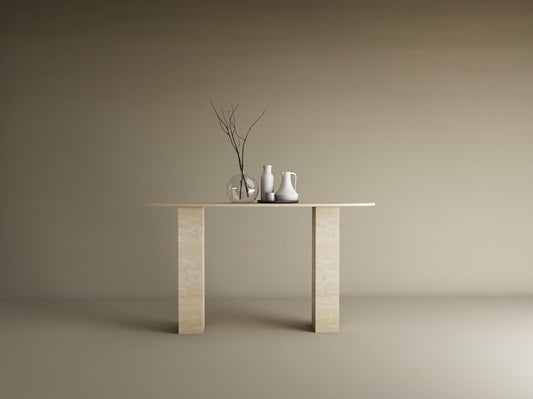 The TRAVERTABLE Ellips Travertine Console Table with a sculptural rectangular base displays three vases—one clear, two white—with branches in the clear vase, set against a plain beige background.