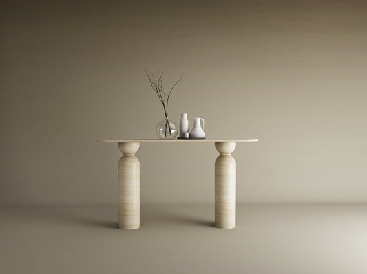 The Ovale Travertine Console Table by TRAVERTABLE, featuring a sculptural round base in organic travertine stone, displays a thin branch in a glass vase, a white pitcher, and jug against a beige backdrop—an understated statement piece.